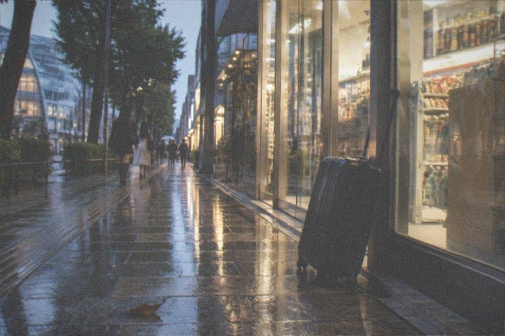 A Tokyo street corner at dusk with a convenience store entrance and a suitcase leaning against the glass.