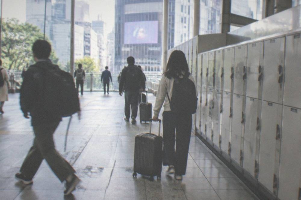 A busy Tokyo station concourse with travellers carrying backpacks and suitcases past a locker bank