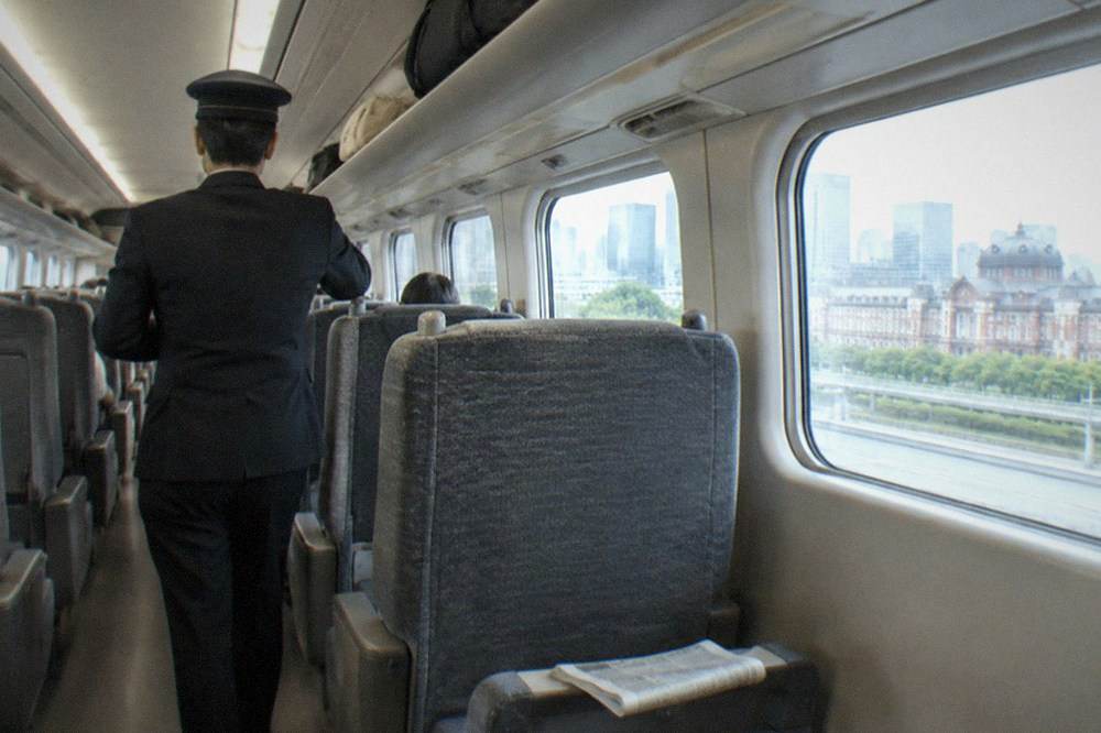 Train conductor standing in the aisle of a Shinkansen carriage beside overhead luggage racks