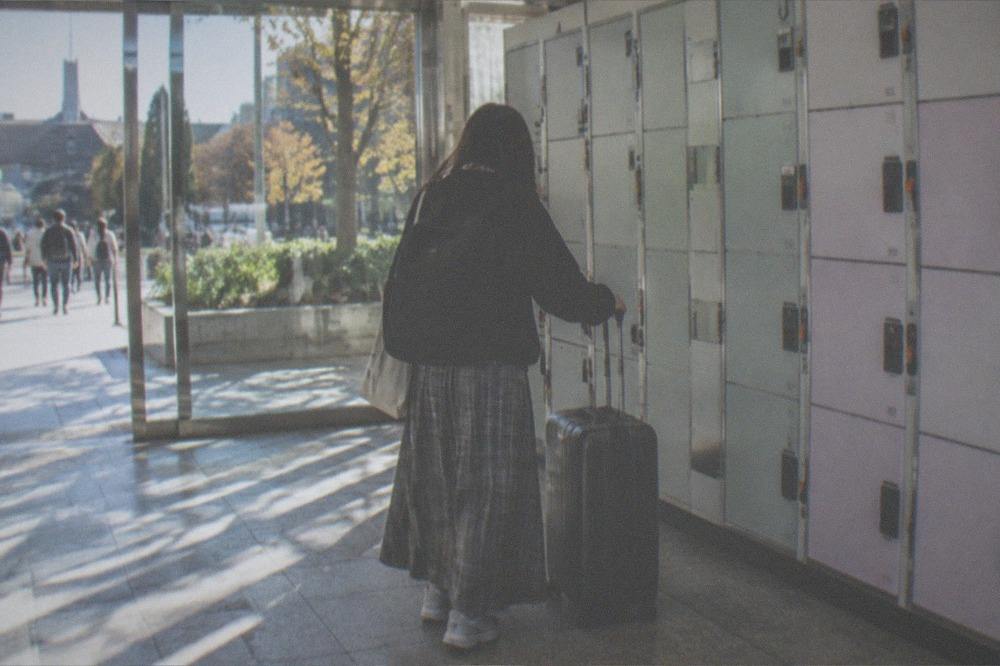 A traveller comparing a suitcase against a row of lockers of different sizes in a station hallway
